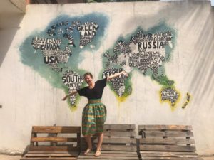 This is a picture of me, Chelsea Bee, standing on a wooden bench with my arms stretched. I am in front of a wall with a painting of the world on it. The countries are black words surrounded by blue, green, and yellow. I am wearing a black shirt and a multi-colored skirt. 