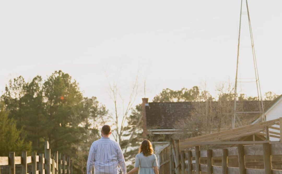 This is a photo of Chelsea Bee and her husband. They are on a path between two wooden fences, facing a barn and windmill. He is wearing a striped shirt and blue jeans, and she is wearing a blue dress and brown cowgirl boots. They are holding hands, and the photo is taken of their backs.