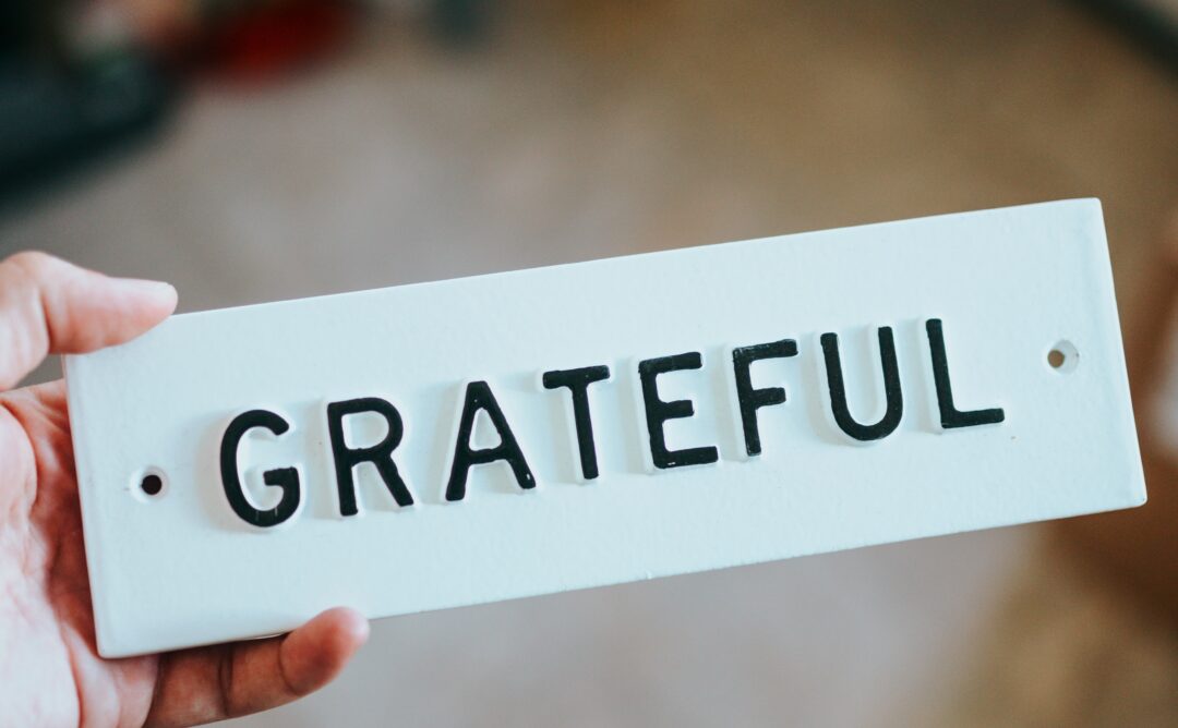 This is a picture of someone holding a white rectangular sign that says “grateful” in black capital letters. All you see is one of the person’s hands.
