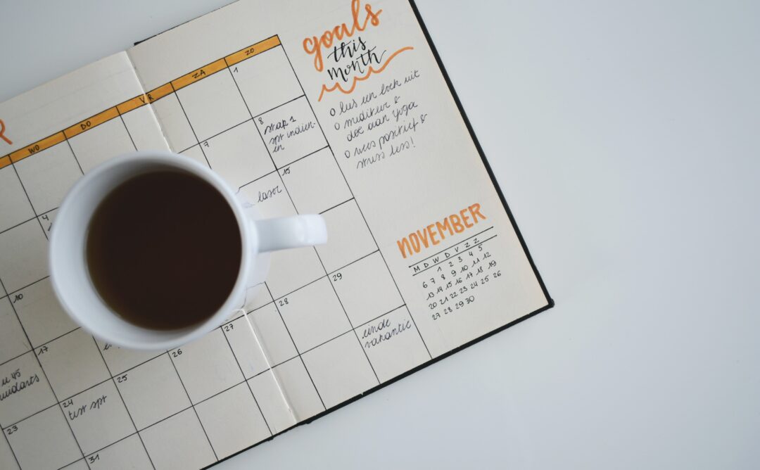This is a picture of a planner open to the November calendar. It is handwritten, and several days have events scheduled. Coffee in a white mug is sitting on top.