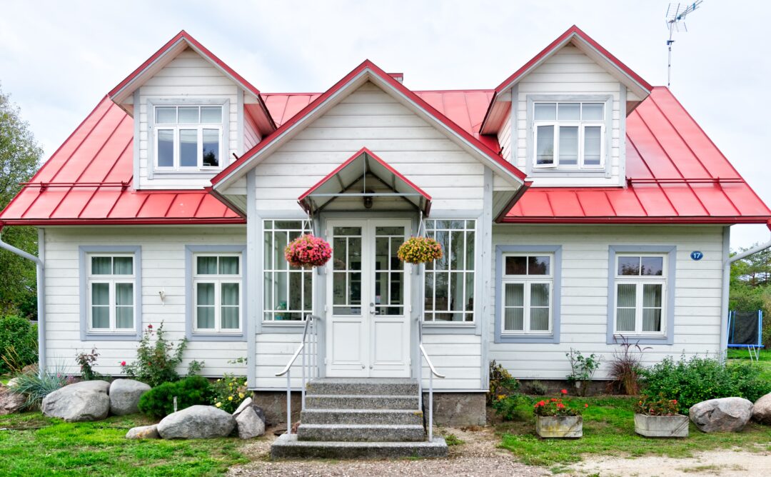 This is a picture of a house with white wooden siding and a red metal roof. It likely has a strong foundation as it is made of rock. The house has two hanging baskets of flowers, one on each side of the door, and has various plants and rocks in front.
