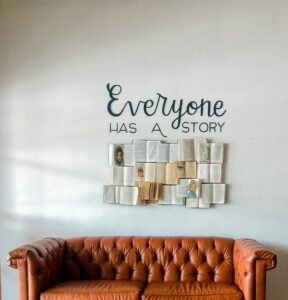 This is a picture of a rust-colored couch, and above the couch multiple books are arranged in a rectangle. Some of the books are open, some are closed, and some have words or drawings on them. Each of the books is a tan or white color. Above the books it says “Everyone has a story” in black font. 