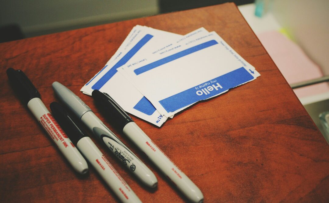 This is a picture of blue and white labels that say “Hello” lying on a brown wooden surface next to 3 black and 1 silver marker. These represent the labels we wear that, overtime, begin to be our identity.