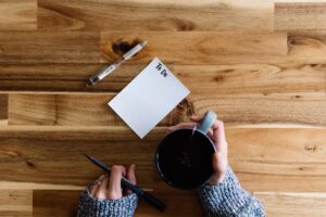 This is a picture of a To Do list on a white note pad with a pen beside it. There is a person’s hands, the left holding a pen and the right holding a coffee mug. Everything is on a wooden surface. This picture represents taking the time to go through the process of leaving your labels at the door.