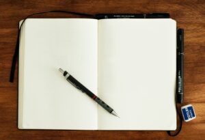 This is a picture of a journal open to two blank pages. There is a black pen on top of the journal. The journal is lying on a wooden surface.