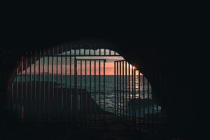 This is a picture of a rock wall with an opening that has a metal, barred gate in front of it. The gate’s door is open. On the other side, you see the dark blue ocean and an orange sky. This represents getting out of the comparison trap.