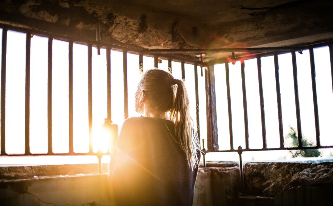 This is a picture of a woman looking out of bars. The ceiling and the wall under the bars are concrete. The woman is facing away from the camera and she is holding onto the bars. Sun is shining through.