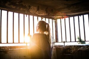 This is a picture of a woman looking out of bars. The ceiling and the wall under the bars are concrete. The woman is facing away from the camera and she is holding onto the bars. Sun is shining through.