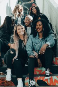 This is a group of 6 women sitting on stairs. They are women of various skin and hair colors. All of the women are smiling, some looking at the camera and some looking at each other. 