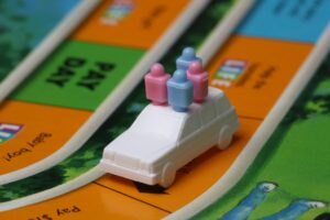 This is a white plastic car with 4 plastic people in it. Two are pink and two are blue. They are on a orange, blue, and green game-board road.