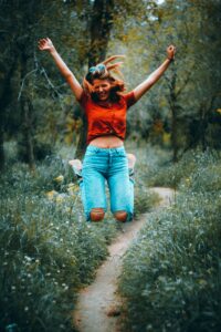 This is a picture of almost woman jumping up into the air. Her legs are bent and her arms are up. She is wearing a red shirt and blue jeans. In the background is a narrow dirt path with green plants and trees on both sides.