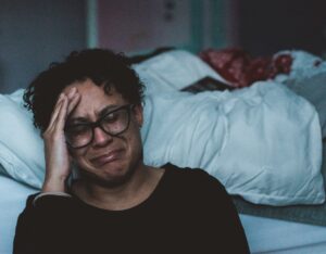 This photo is of a black woman wearing a black shirt and glasses sitting on the floor by an unmade bed with a white comforter on it. The woman has her hand on her head and appears to be crying. This represents the idea that if we don't choose to listen to our bodies, we may eventually be forced to.
