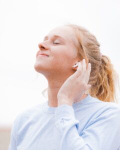 This is a picture of a white woman with blonde hair wearing a light blue shirt. She has her eyes closed and is listening to something through her earbud. Her hand is on her ear. This photo represents listening to your body.