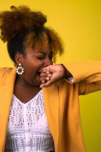 This is a picture of a black woman wearing a white shirt and a golden yellow blazer. She is yawning and has her hand over her mouth. The background is yellow. Fatigue is one thing our body might be using to speak to us.