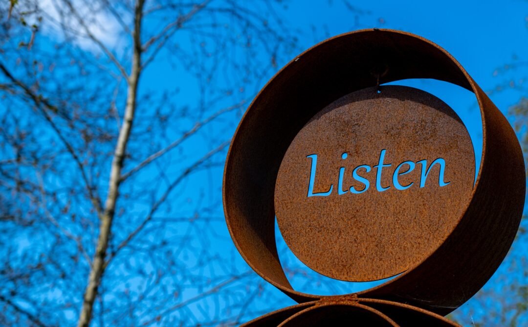 This is a picture of a rusted, circular metal sign that says "Listen." The background is a blue sky with a brown tree with no leaves. It represents the main idea of the blog post, "Are You Listening to Your Body?"