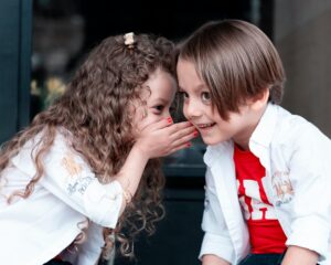 This picture is of a little girl with curly brown hair telling a little boy a secret. The boy has straight brown hair and is smiling. Both of the children appear to be white. 
