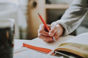 This is a picture of a hand writing in a cream-colored notebook with a red pen. 