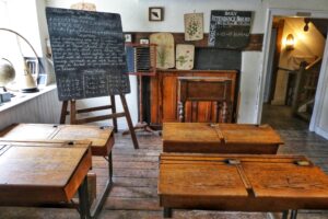 This is a photo of an old-timey school classroom. There are several wooden desks in two rows. At the front, there is a bookcase, various pictures, and a blackboard on a stand with white writing on it. It represents lessons I have learned from marriage.