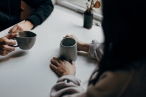 This is a photo of two people sitting at a table drinking coffee out of gray pottery mugs. The person on the right has long black hair. It represents both talking to others about hard things and not being alone in your struggles.