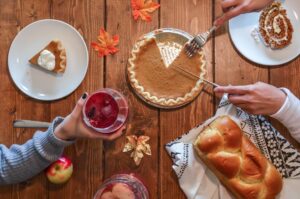 This is a picture of, what appears to be, a Thanksgiving meal. There is pumpkin pie on the right side with one slice cut out of it. Someone has a fork and knife held above the pie. Another person on the left is holding a glass with a pinkish-purple drink in it. In the lower right corner there is a loaf of bread on a white towel. The food is on a wooden surface with multi-colored leaves scattered around. 