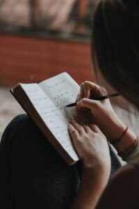This is a picture of a person, writing in a notebook. The paper is white and the ink is black. You mainly see the person’s hands, but he or she does have black hair and black pants. 