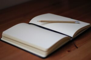 This is a picture of an open journal. It shows 2 white pages, a black ribbon bookmark in the middle, and a pencil on the right side. Journaling is a way to reflect about living an intentional life. 