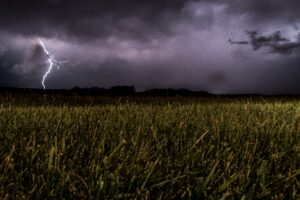 This is a picture of a storm. The sky is dark gray and black and covered with clouds. There is also a strike of lightning on the left side. The field of grass is also dark and in shadows. 