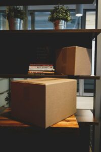 This is a picture of shelves in a room. They hold 2 plants on the top, a stack of books and a brown box in the middle, and a brown box on the bottom. The boxes represent the “40 Bags in 40 Days Challenge” I am going to complete. This is part of my way of observing Lent. 