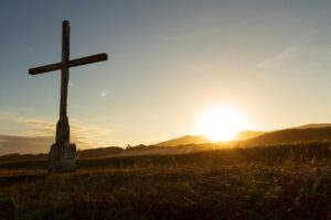 This is a picture of a cross in a field. The sun is low in the sky and the picture is a variety of golden tones. 