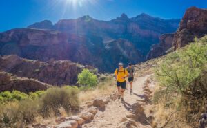 This a picture of a man and a woman running on a dirt trail. There are green plants on either side of the trail, mountains are in the background, and the sky is blue.