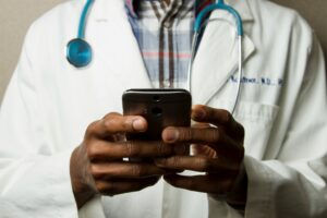 This is a picture of a doctor wearing a white lab coat over a plaid shirt. He has a stethoscope around his neck and is holding a cell phone in both hands. The picture only shows the man’s upper body, starting at the top of his shoulders.