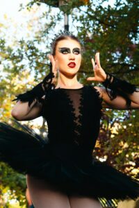 This is picture of a woman wearing a Black Swan costume, including a black leotard with tutu, a crown, dark eye makeup, and red lipstick. She is outside with trees behind her. Black Swan is one of the movies about women and mental health.