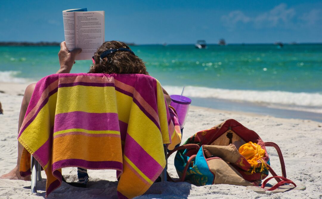 This is a picture of a person sitting on the beach in a chair reading a book. The photo is taken from behind the person, and they are facing the ocean. There is a yellow, pink, and orange striped towel on the back of the chair. There is a bag in the sand beside the chair.