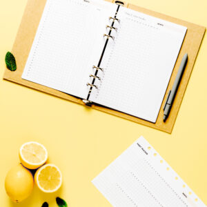 This is a picture of a yellow binder open to habit tracking pages. There is a gray pen on the right side. In the bottom right corner there is another habit-tracking page. In the bottom left corner there are lemons, one whole and one cut in half. The background is light yellow. This picture represents adding structure into my life when I transition from summer break to work.