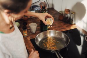 This is a picture of a woman cooking. She is pouring something from a small cup into a frying pan on a stove. The kitchen counter is in the background, and there are several cooking items on it. 