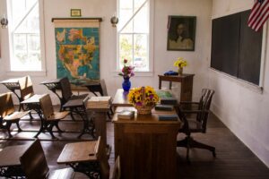 This is a picture of an empty school classroom. There is a teacher’s desk and chair on the right side of the photo and several student desks on the left. There are flowers and books on the teachers desk. On the walls in the background, there is a framed photo of a woman and a map of the United States. There is also a window. This photo can represent the transition from school to summer or the transition from summer to school.