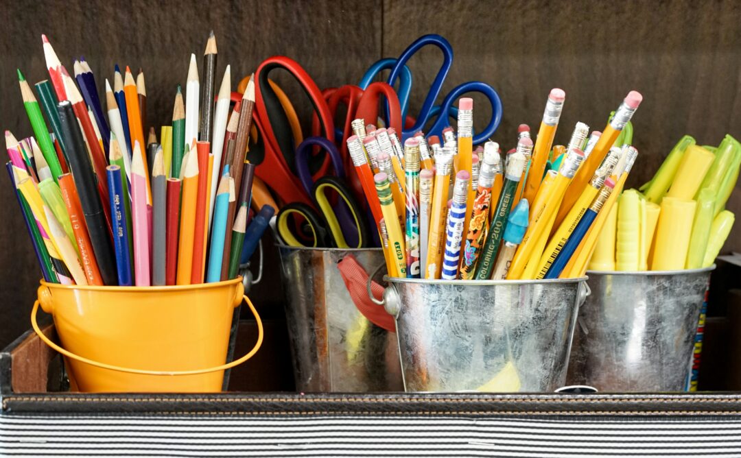 This is picture of various school supplies in metal buckets. Starting from the left: colored pencils in a yellow bucket, scissors in a gray bucket, pencils in a gray bucket, and highlighters in a gray bucket.