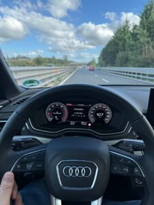 This is a photo from the driver’s point of view in car. You see the steering wheel and the road ahead. There are green trees on the right side of the road and a blue sky with white clouds on the left.