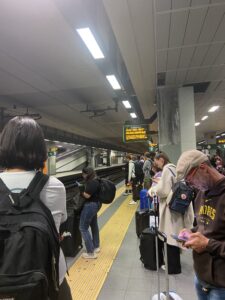 This is a picture I took while we were waiting in an underground train station. There are a variety of people, most with a backpack or suitcase. The train track is on the left, but isn’t very visible due the people standing beside it. In the middle, there is a yellow line on the floor and a row of lights on the ceiling. There is also a sign in the top middle of picture with orange words on it. 