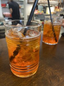 This is a picture of two orange aperol spritzes. They are in clear glasses with black straws sitting in a wooden table. Each of the drinks has ice in it. 