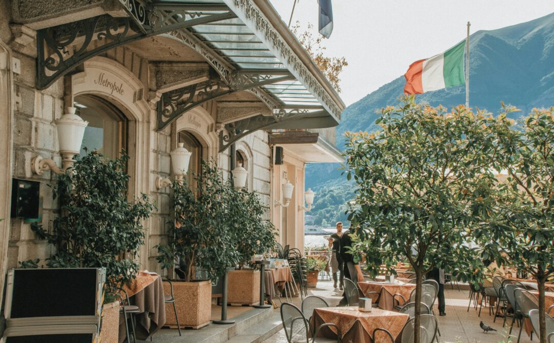 This is a picture taken in a town in Italy. It is outside a light brown, stone building. There are tables and chairs on the right side of the picture. There are small trees beside the building and among the tables. In the top right corner is a red, white, and green Italian flag.