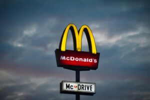 This is a picture of a yellow and red McDonalds sign against a dark gray-blue, cloudy sky. The yellow M is at the top with the red sign that says “McDonalds” in white capital letters is underneath. At the bottom, there is a smaller white sign that says “McDrive” in black capital letters.