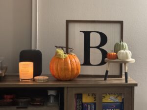 This is a photo of fall decor on top of a wooden cabinet. On the left side, there is a lit candle, glowing orange in a jar. The wooden top of the jar is sitting beside it. Beside the candle is an orange pumpkin, a white wooden square sign with a black B on it, and 3 small pumpkins on a small wooden stool. The pumpkins are orange, sage, and white. The sign is sitting slightly behind the pumpkins.