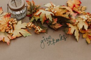 This is a picture of multiple orange and brown leaves that forms an arch on a brown paper background. In the top left corner there is a glass and rusted metal lantern. In the middle right “give thanks” is written in black cursive.