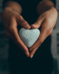 This is a picture of a brown hands holding a gray heart-shaped rock that says “Hope” on it.