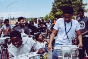 This is a picture of numerous black children riding bikes in a group. In the front row (left to right) there is a boy wearing a gray sweatshirt who is smiling, a younger girl in a white shirt, and a boy with a white shirt and a blue lanyard. Behind these three there are many more kids and teens.