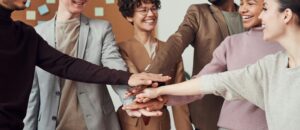 This is a picture of 7 people, both male and female, of different skin colors or nationalities. They are all standing side by side with their hands stretched to the middle making a stack. The faces you can see are smiling.