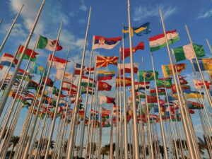 This is a picture of numerous flags from different countries, each on its own flagpole. In the background you see blue sky and thin white clouds.